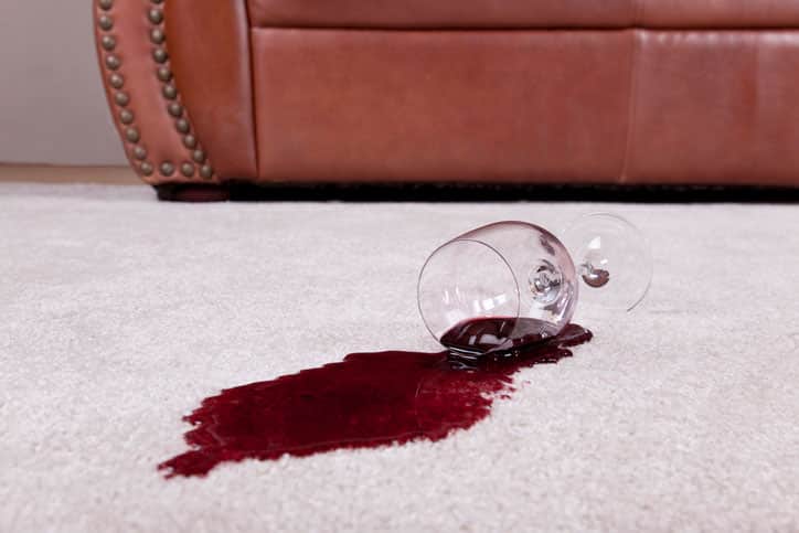 A wine glass with a red wine spill creating a puddle rests on a tan loop pile carpet. The wine glass rests on the spilled wine. The glass has a long stem and wide base. The spill fans out from the top of the glass. The spill appears deeper underneath the glass and then becomes a little lighter as it fans out. The spill is oval in shape. A tan leather couch with decorative metal buttons that run up the side sits in the background. Neutral colored walls are also in the background.